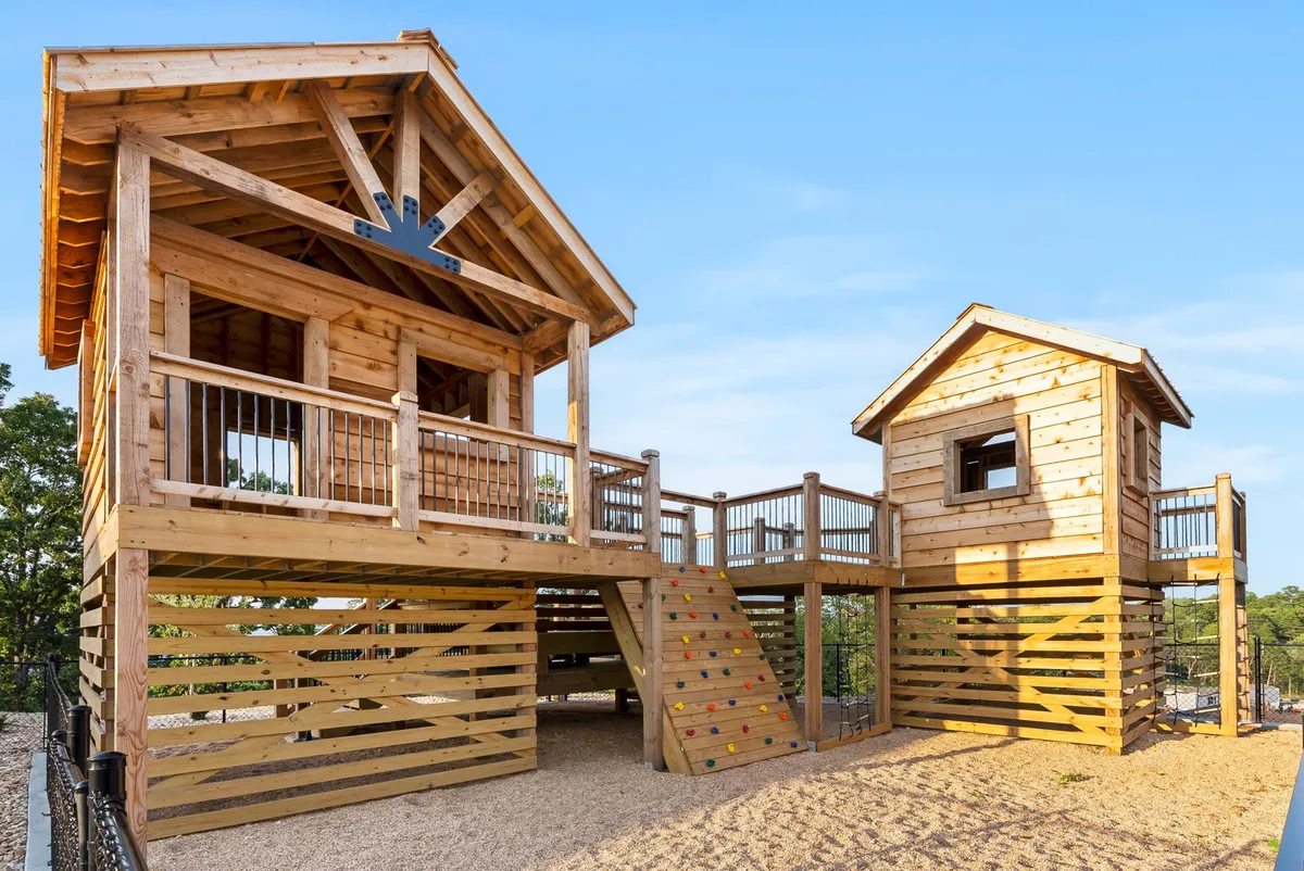 A colorful exterior view of an adventurous playhouse structure with climbing walls, a slide, and children actively exploring and playing among the surrounding trees. The setting is joyful and brimming with imaginative activity, ideal for younger guests.