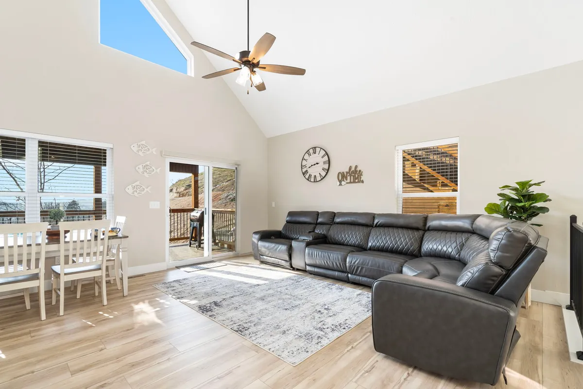 A spacious, open-concept interior showcasing a large sectional sofa, a dining table, a sliding glass door leading to the deck, a decorative wall clock, and a plush area rug. Natural light streams in, creating a cozy and inviting atmosphere ideal for gatherings, meals, and relaxation.