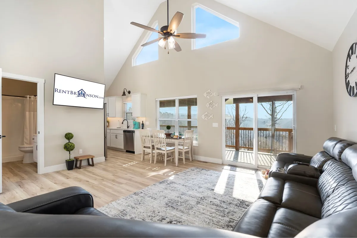 The image showcases a spacious, light-filled interior with large windows overlooking a private wooded view. The inviting living space includes a modern kitchen, a welcoming dining table set for a group, and a cozy seating area perfect for relaxing or gathering with family. Warm natural light floods the space, highlighting the connection to the outdoors and the tranquil setting.