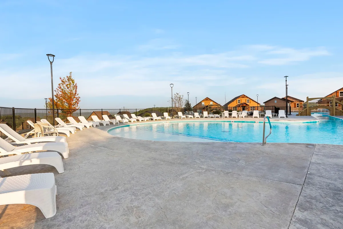 A sun-soaked swimming pool with ample lounge chairs sits at the heart of the resort, with surrounding cabins visible in the background. The pool area is lively yet relaxed, inviting guests of all ages to swim, sunbathe, or gather with friends and family for a fun group experience.