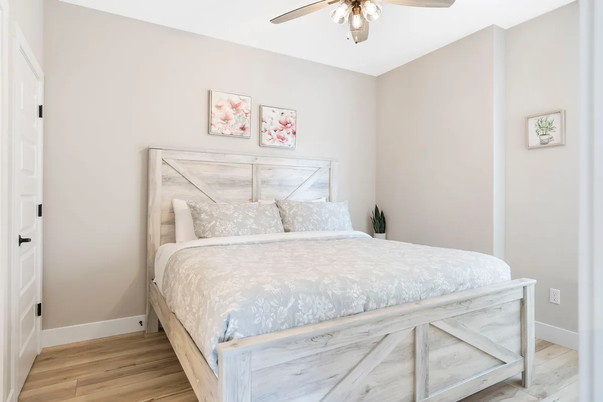 A serene king-sized bedroom with a large, comfortable bed adorned with plush linens, decorative wall art, and a ceiling fan. Soft morning light filters in, creating a calm, peaceful atmosphere perfect for unwinding after a busy day.
