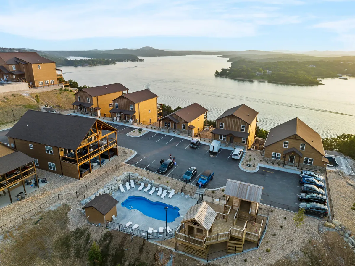 This exterior image captures the central swimming pool framed by wooden cabins, a scenic lake view in the background, and a vibrant playground area. There are families relaxing by the pool, children playing, and the atmosphere is lively yet relaxed, embodying both recreation and natural beauty.