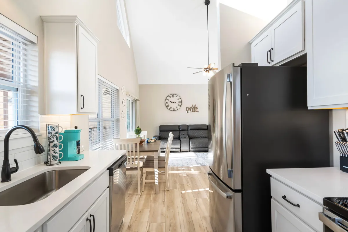 A cozy, open-plan living space combining a kitchen sink area, dining table, and living room couch, centered around a large wall clock. The room is designed for cooking, dining, relaxing, and socializing, with a cohesive, welcoming atmosphere.