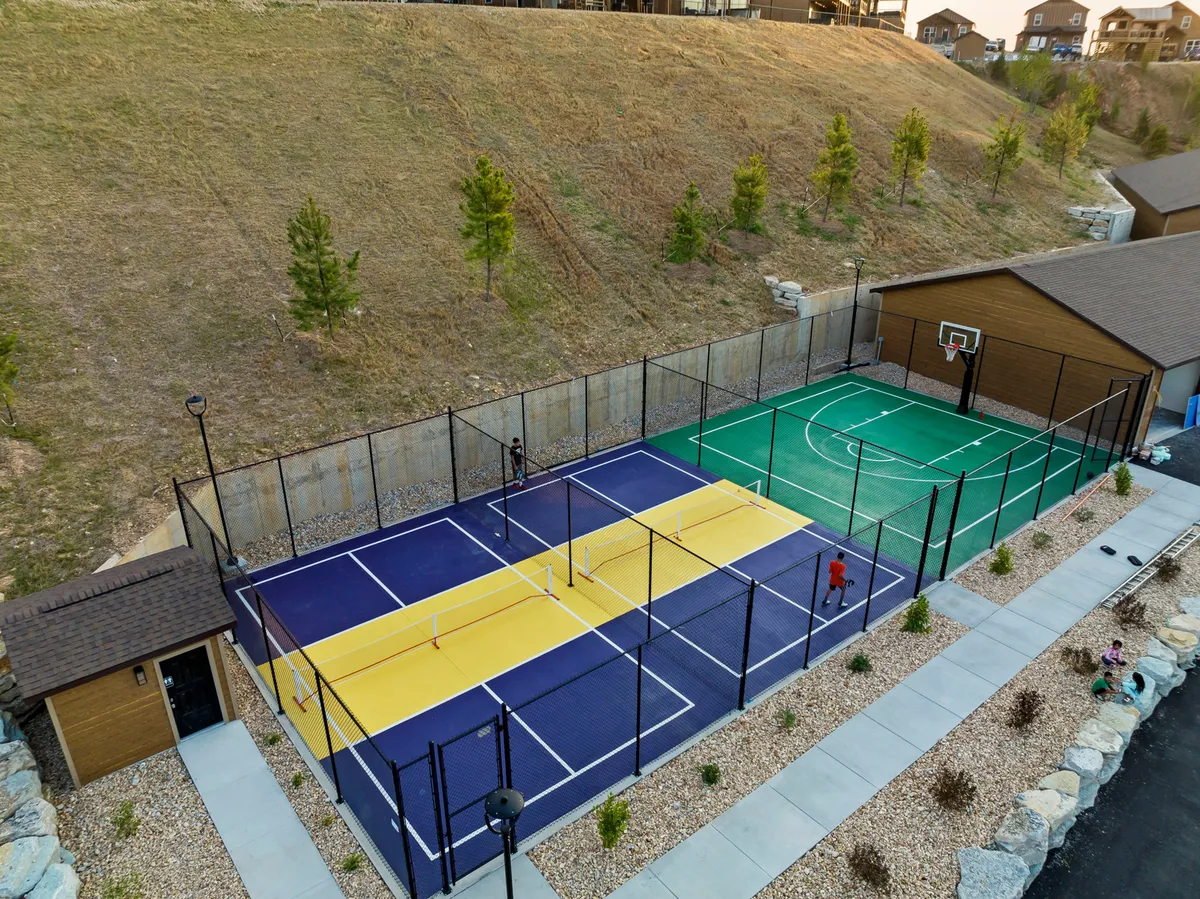 An energetic exterior view of a multi-sport court set amidst lush greenery, featuring a basketball hoop and open space for various activities. People are playing and socializing, adding a dynamic, active vibe.